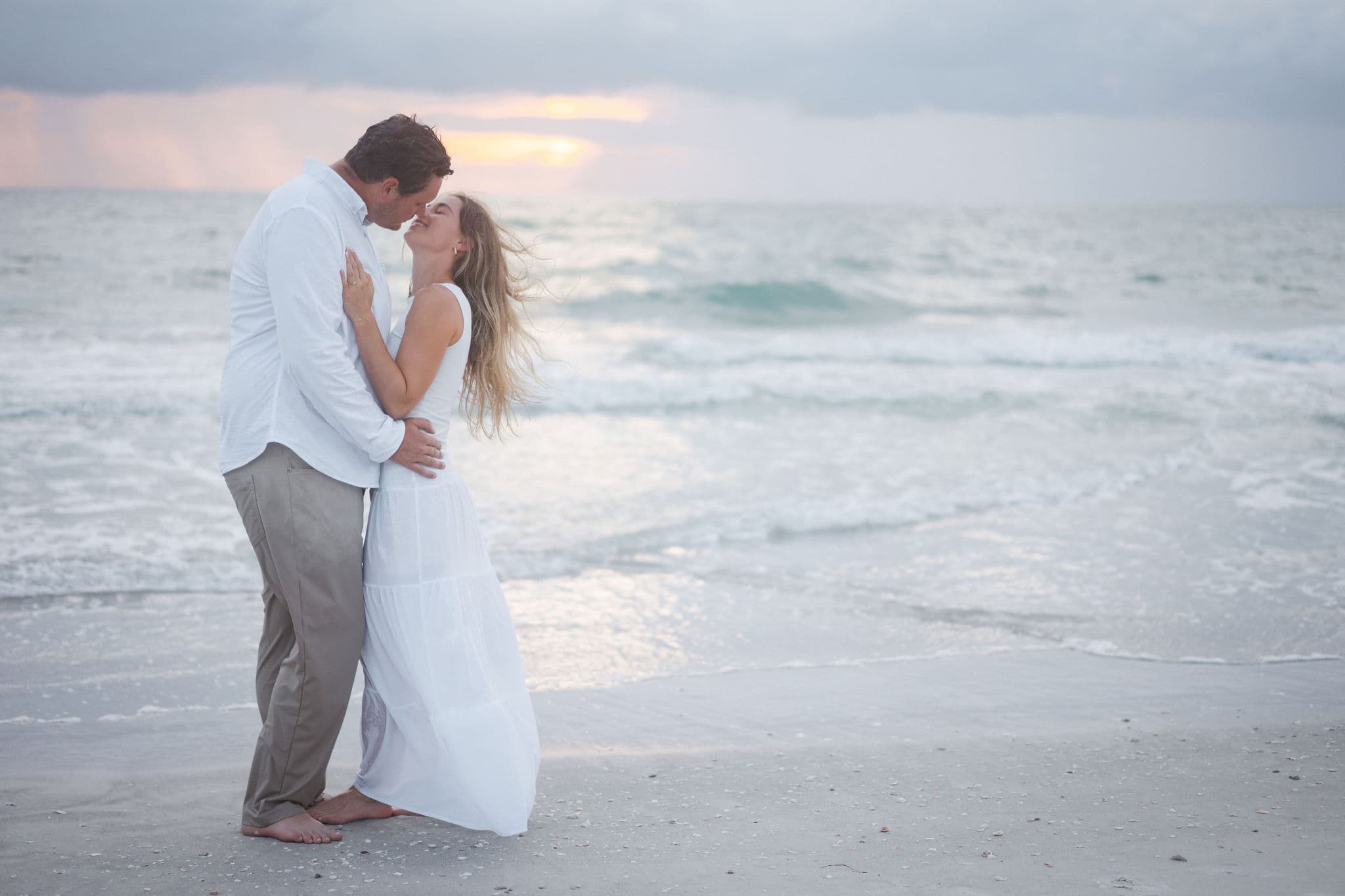 Couple nuzzling at the shoreline under a moody peach sunset sky