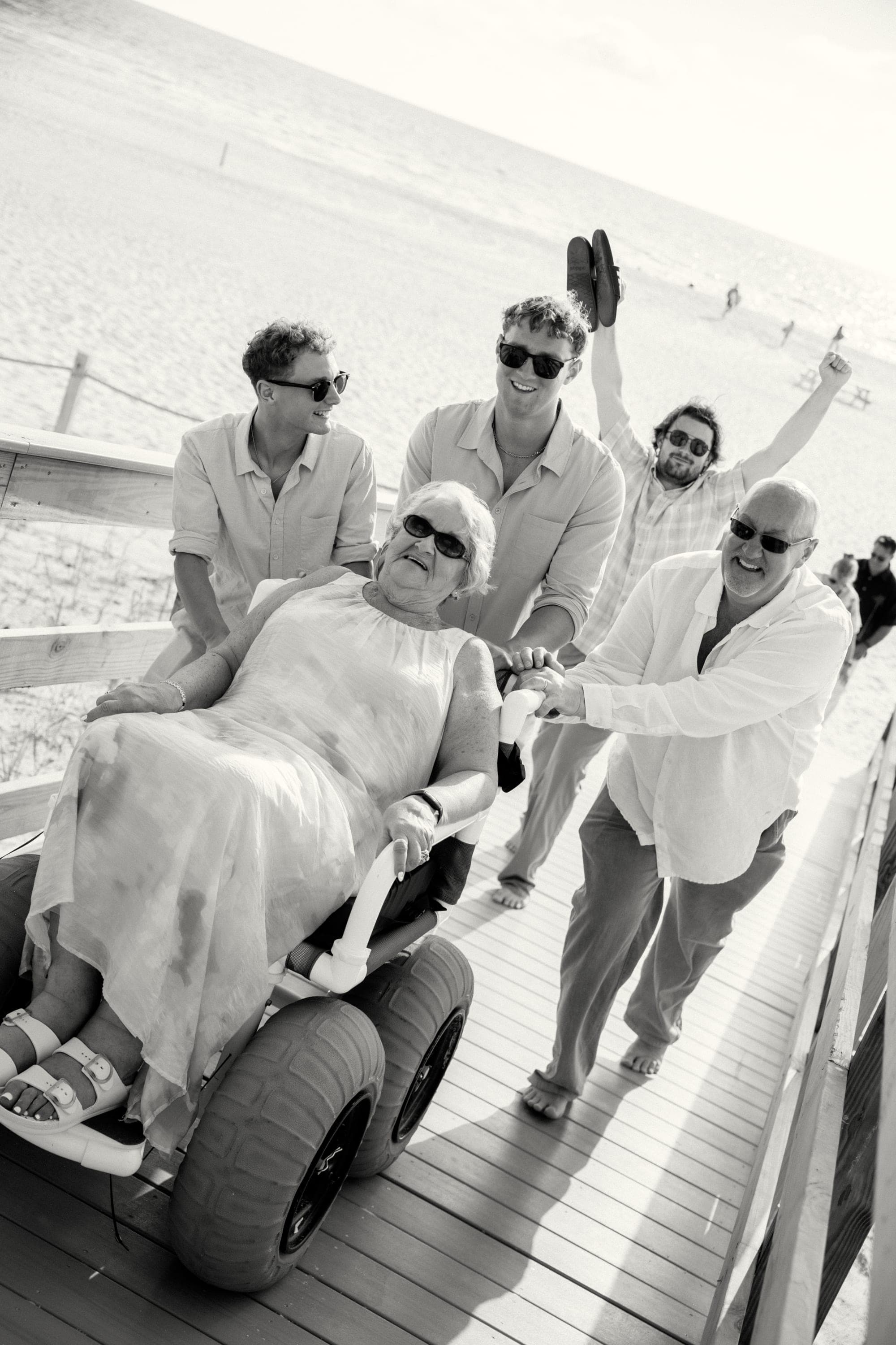 Family joyfully pushing a smiling grandmother down a beach boardwalk, black and white