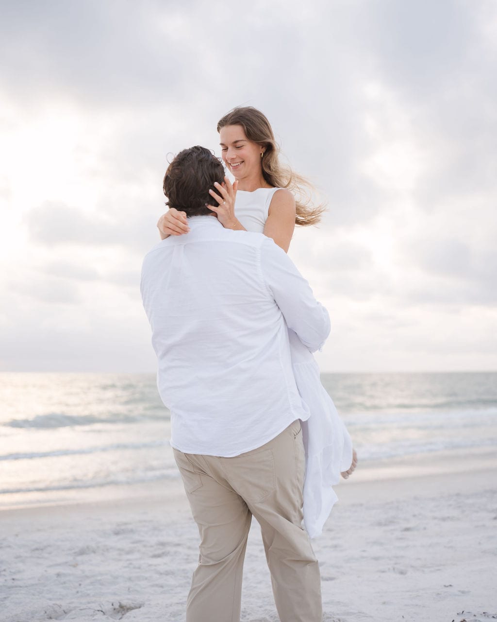 Fiance lifting their partner, both in white outfits, on a Gulf coast beach