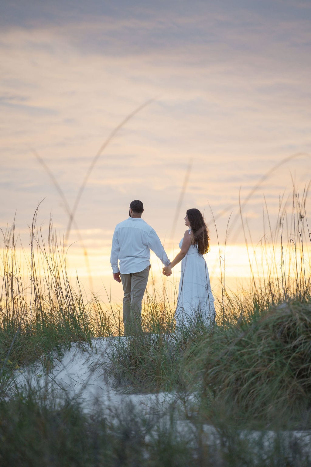 Couple holding hands walking through beach dunes at golden sunset in Sarasota