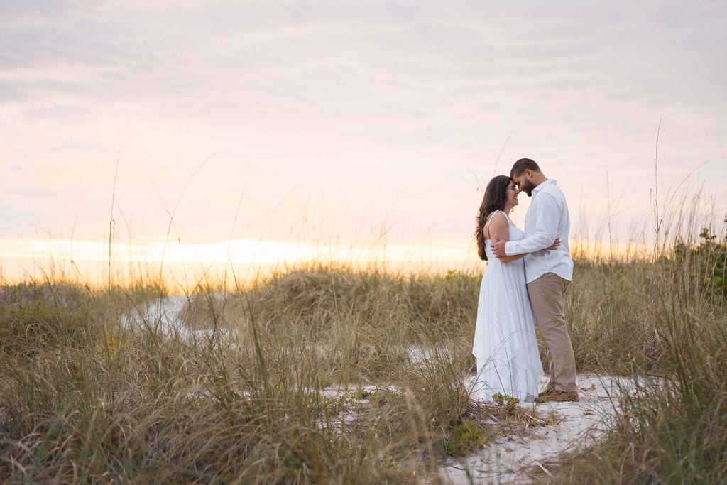 Couple with foreheads together among beach dunes and sea oats at sunset