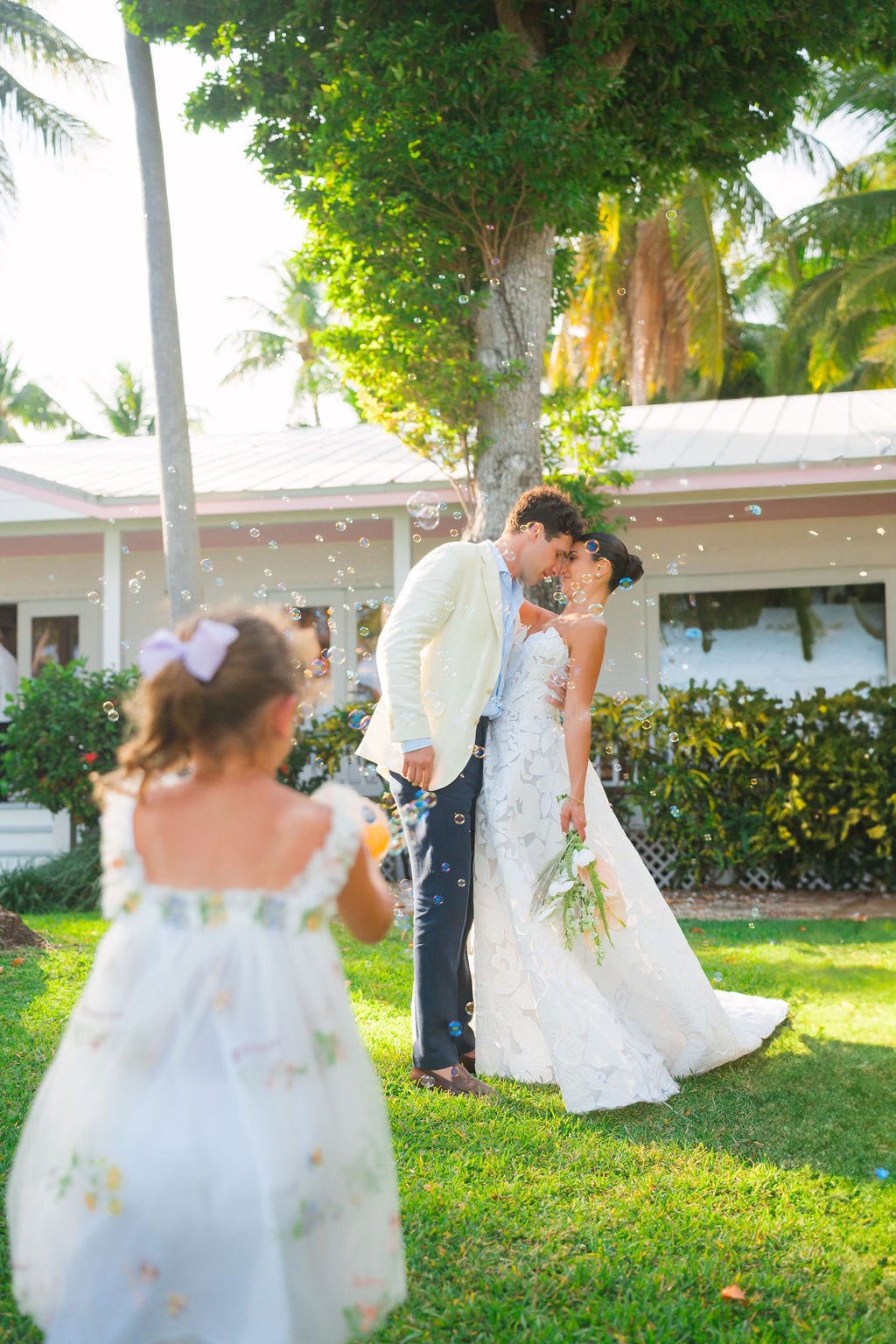 Bride and groom touching foreheads while a flower girl blows bubbles nearby